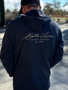 A cozy Bella Luna Estate Winery Unisex full zip hoodie in a stylish District Brand, crafted from 100% recycled fabric – a blend of 60% recycled cotton and 40% recycled polyester. The hoodie is of ideal weight, featuring a unique Bella Luna doodle by Sherman on the front left chest, the iconic Bella Luna Estate Winery logo on the back, and coordinates elegantly displayed on the right sleeve cuff. Perfect for both comfort and sustainability, this hoodie is a fashionable choice for wine enthusiasts and eco-conscious individuals alike.